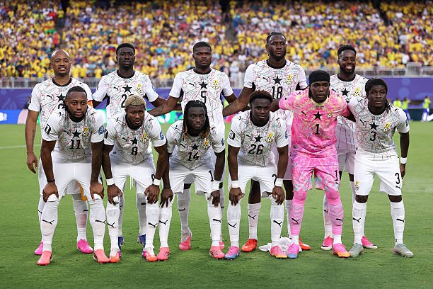 Ghana national football team poses for a pre-match group photo on the pitch, in white kits with black stars; pink goalkeeper stands among teammates as fans cheer in the stands. Ghana national football team poses for a pre-match group photo on the pitch, in white kits with black stars; pink goalkeeper stands among teammates as fans cheer in the stands.