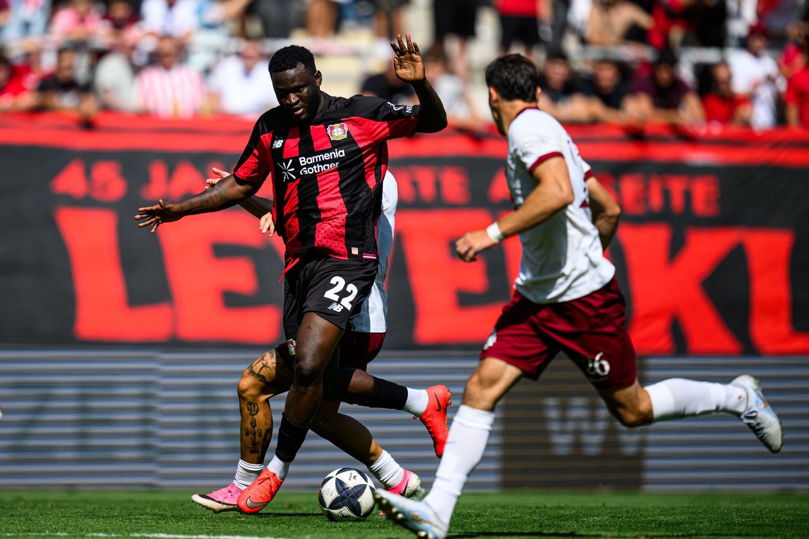 Soccer player in a red-and-black striped jersey dribbles the ball as a white-uniform opponent closes in, with a crowded stadium in the background. Soccer player in a red-and-black striped jersey dribbles the ball as a white-uniform opponent closes in, with a crowded stadium in the background.