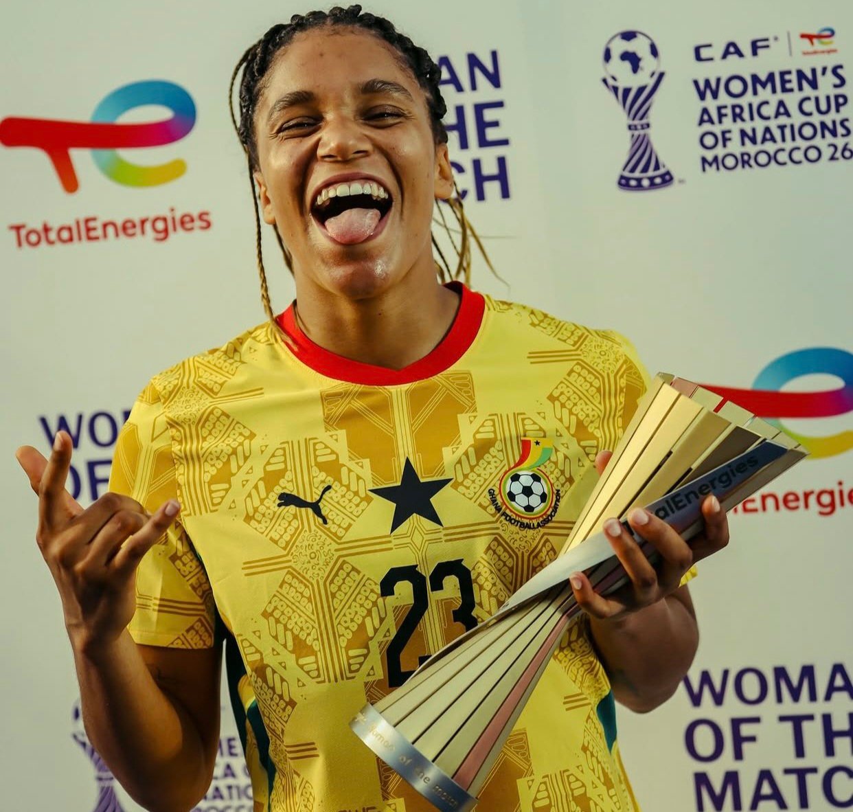 Female soccer player in a yellow jersey (number 23) smiles broadly while holding a multi-panel trophy after a win.