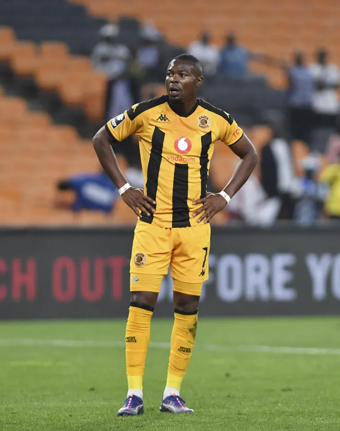 Kaizer Chiefs footballer standing with hands on hips on the field in a yellow and black striped kit.