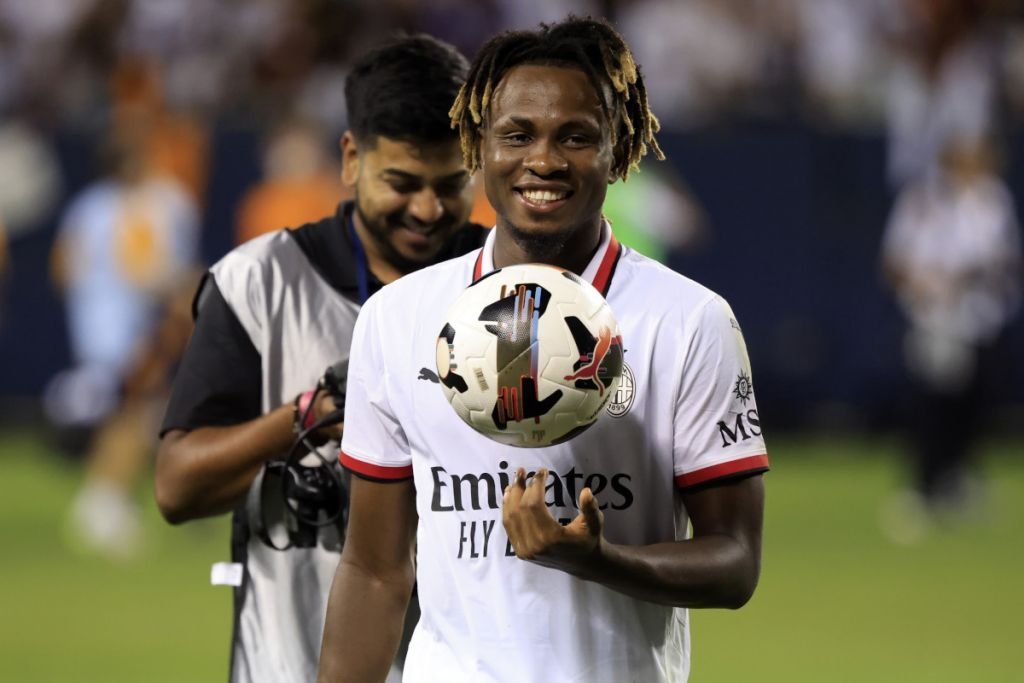 Soccer player in a white Emirates jersey smiling with a ball resting on his chest; photographer in background.