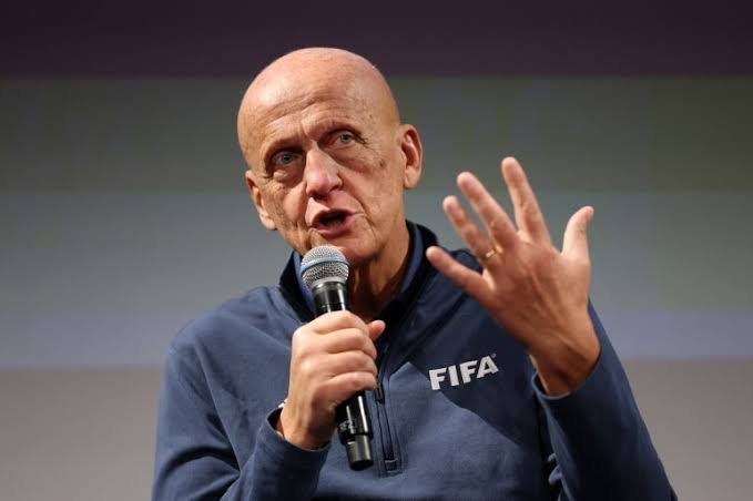 Man wearing a dark blue FIFA jacket speaks into a handheld microphone on stage, gesturing with his left hand. Man wearing a dark blue FIFA jacket speaks into a handheld microphone on stage, gesturing with his left hand.
