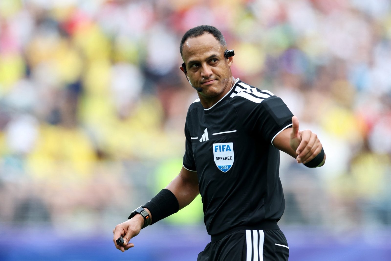 Soccer referee in black kit with a headset, signaling with his right hand on a tense stadium pitch. Soccer referee in black kit with a headset, signaling with his right hand on a tense stadium pitch.