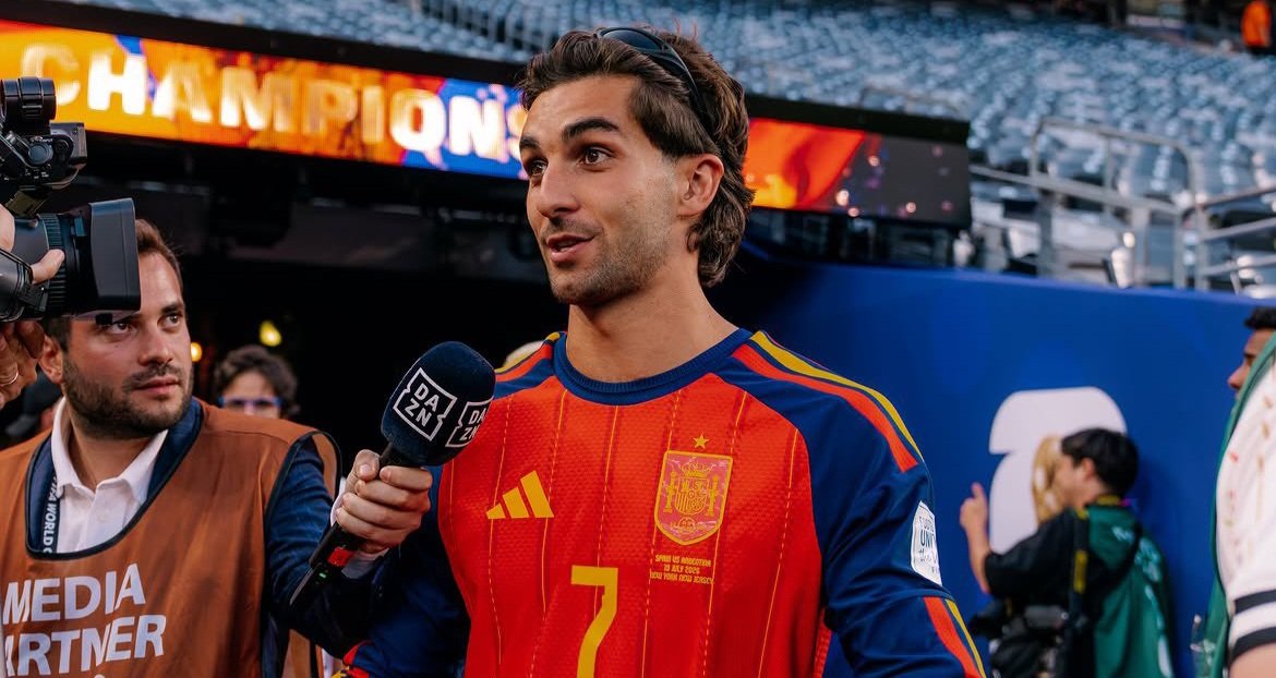 Soccer player in red Spain jersey being interviewed by reporters, microphone labeled DAZN in foreground, Champions banner behind them