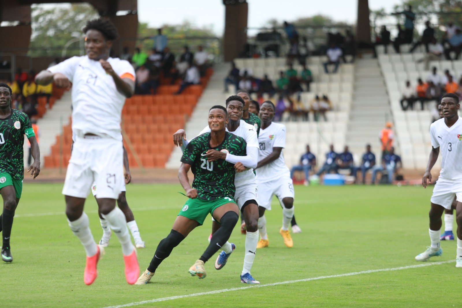 Soccer match action as a green-jersey player (number 20) fights for the ball while a white-jersey defender closes in from behind.