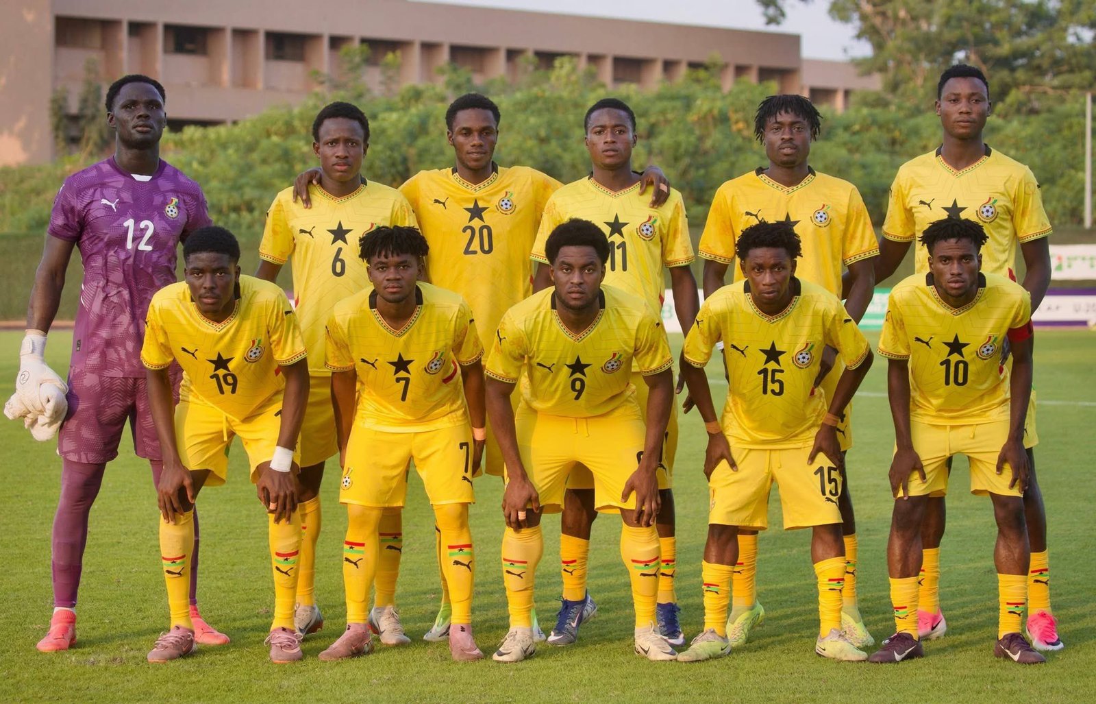 Soccer team in yellow kits posing for a group photo on a grassy field, with a purple-clad goalkeeper on the left.