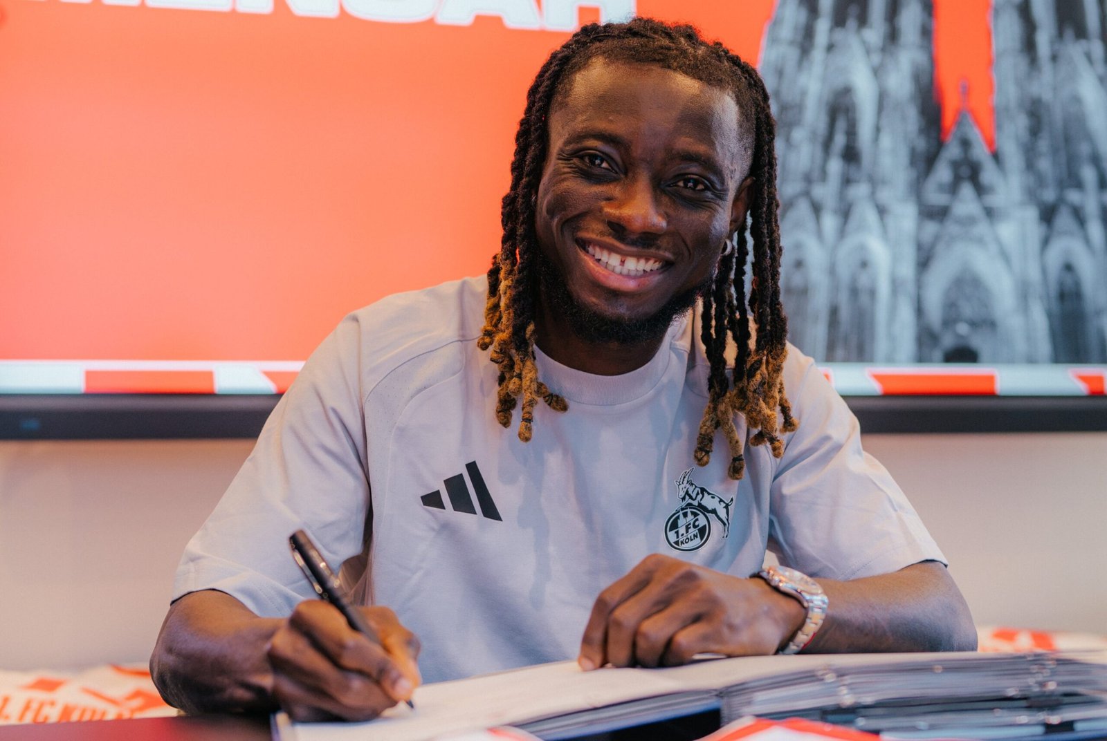 Man with dreadlocks smiling while signing a document at a desk, wearing a light gray shirt with a crest, orange background behind. Man with dreadlocks smiling while signing a document at a desk, wearing a light gray shirt with a crest, orange background behind. Man with dreadlocks smiling while signing a document at a desk, wearing a light gray shirt with a crest, orange background behind. Man with dreadlocks smiling while signing a document at a desk, wearing a light gray shirt with a crest, orange background behind.