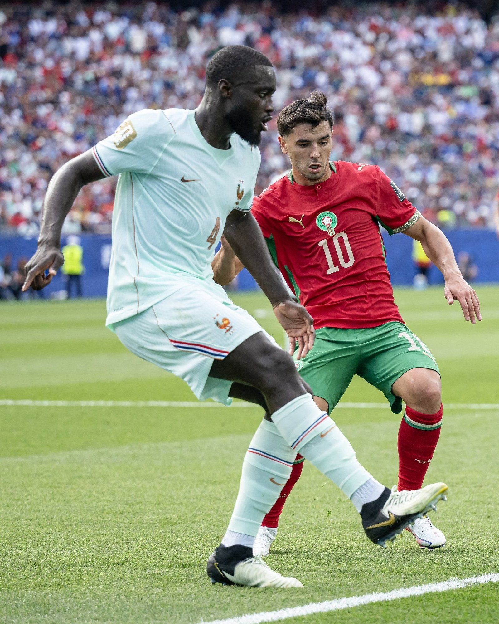 Football match action: a French player in light blue tries to reach the ball while a Moroccan player in red pursues, crowd in the background.