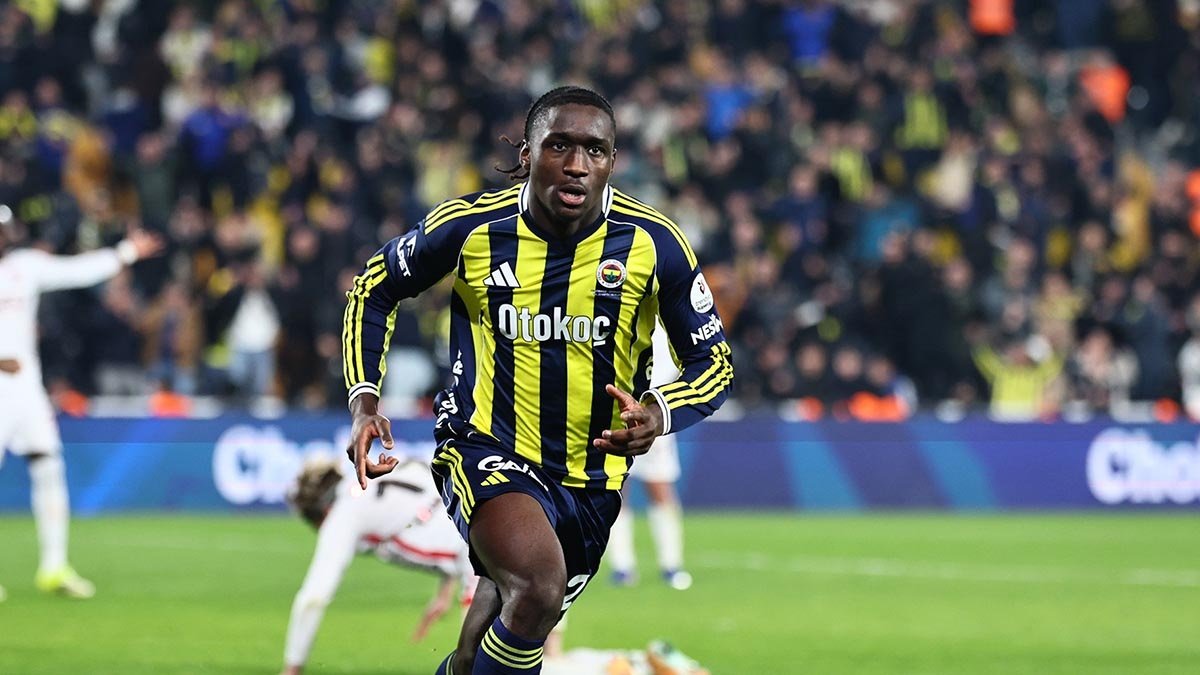 Footballer in yellow and navy striped kit runs on a grass pitch during a match, with a crowded stadium in the background.