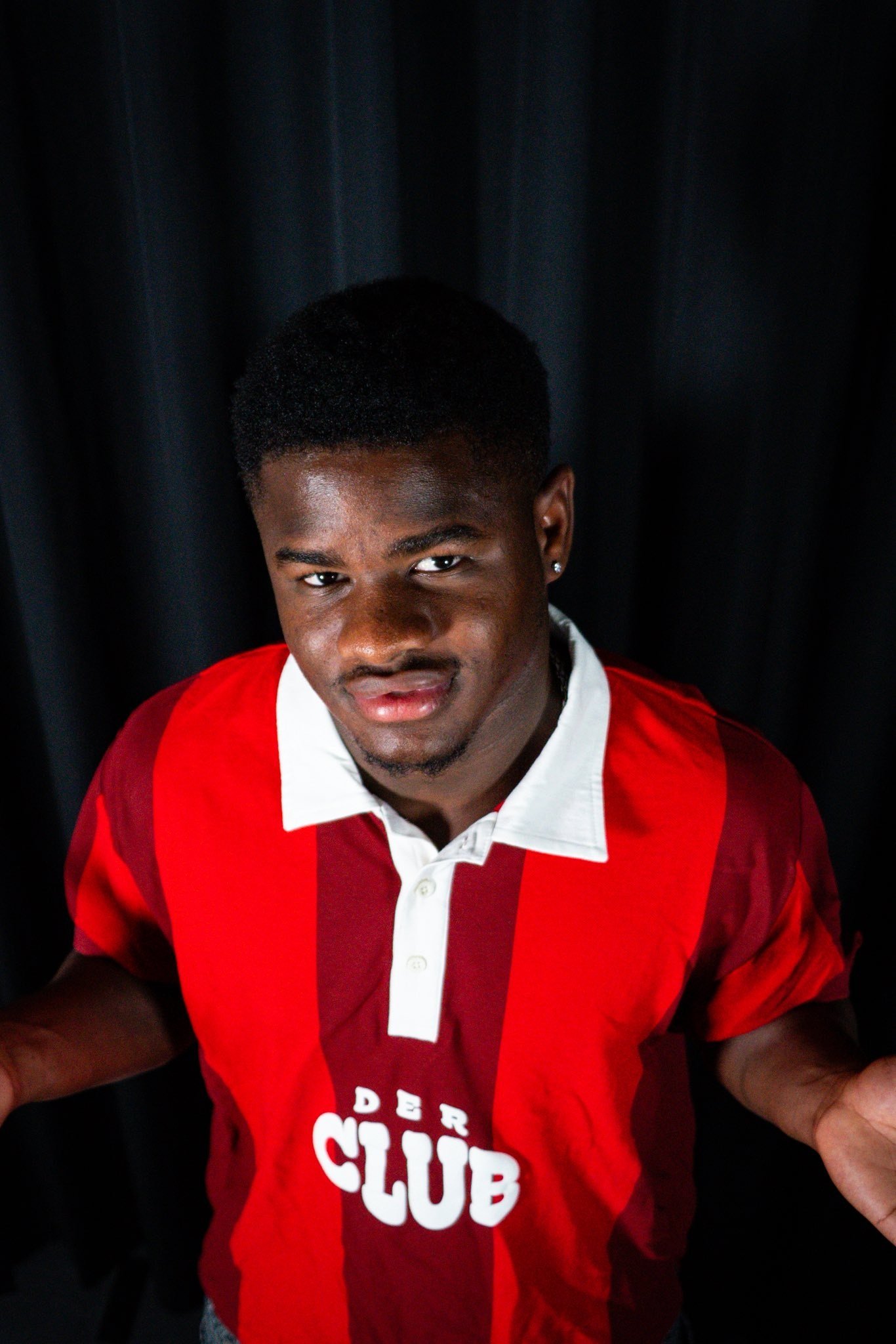 Young man with short hair wearing a red and white striped shirt, standing against a dark backdrop and looking at the camera.