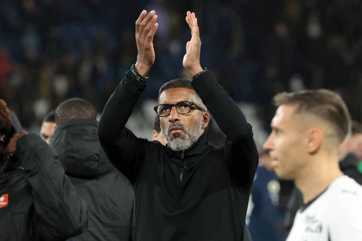 Man with gray beard and glasses raises his hands to applaud on a sideline or field edge. Man with gray beard and glasses raises his hands to applaud on a sideline or field edge.