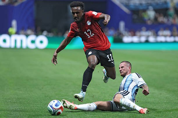Soccer player in red jersey (number 12) jumps while a defender in light blue and white slides in to challenge for the ball on a green pitch. Soccer player in red jersey (number 12) jumps while a defender in light blue and white slides in to challenge for the ball on a green pitch.