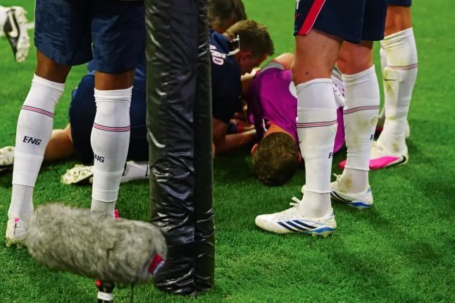 Soccer players in white socks circle an injured teammate lying on the grass while a trainer attends to them.