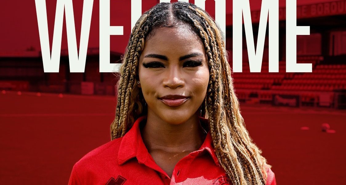 Portrait of a woman with braided hair wearing a red polo, standing in front of a red-tinted stadium with a large 'WELCOME' banner overhead.
