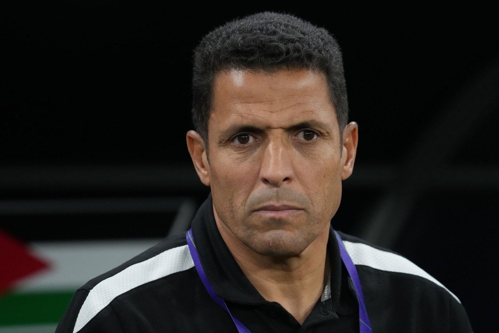 Close-up of a man with short dark hair and a serious expression, wearing a black-and-white polo and a purple lanyard.