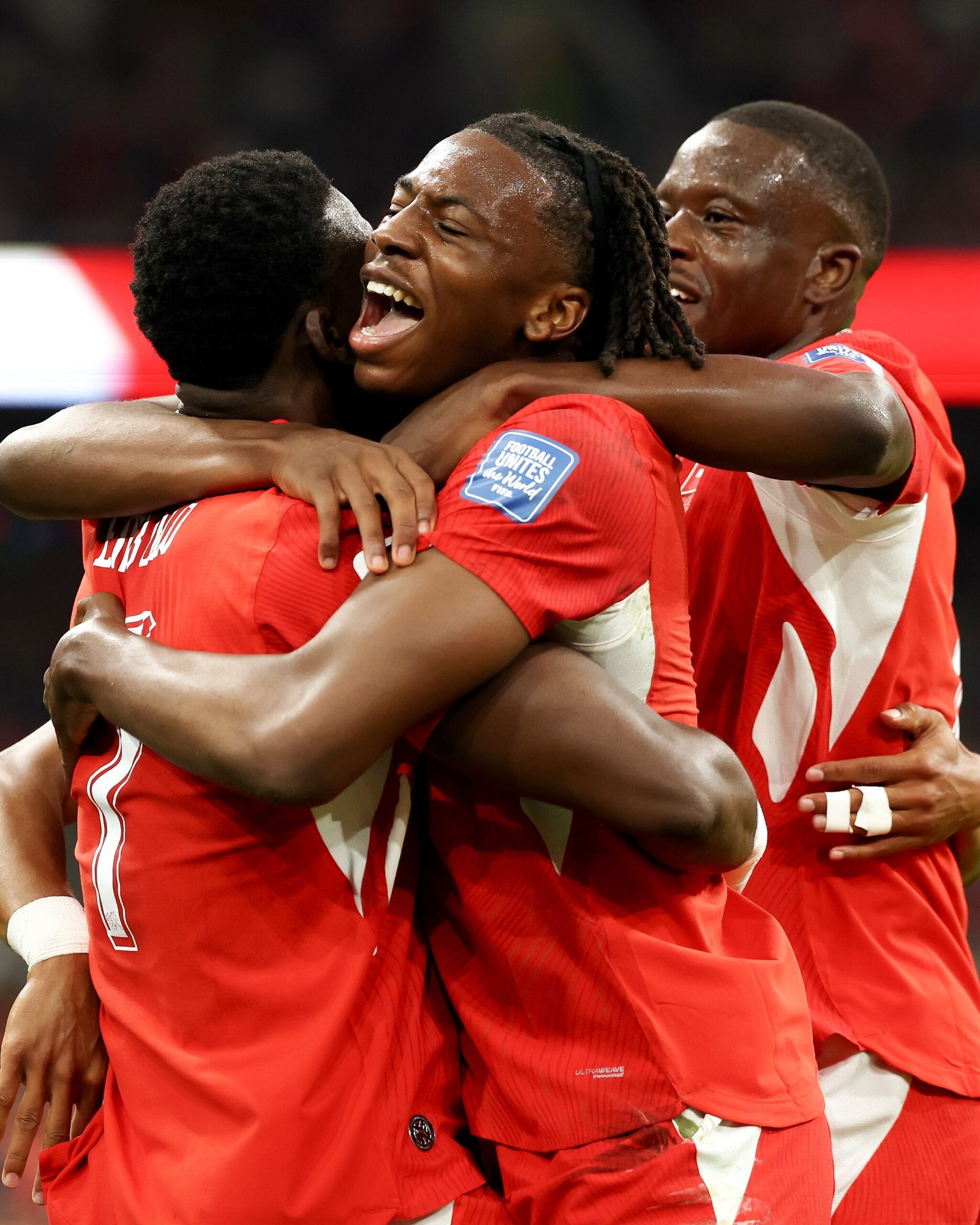 Soccer players in red uniforms embrace in a group hug, celebrating a goal or win.
