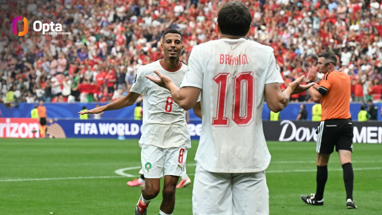 Two soccer players in white jerseys celebrate on a green field as a packed stadium applauds.