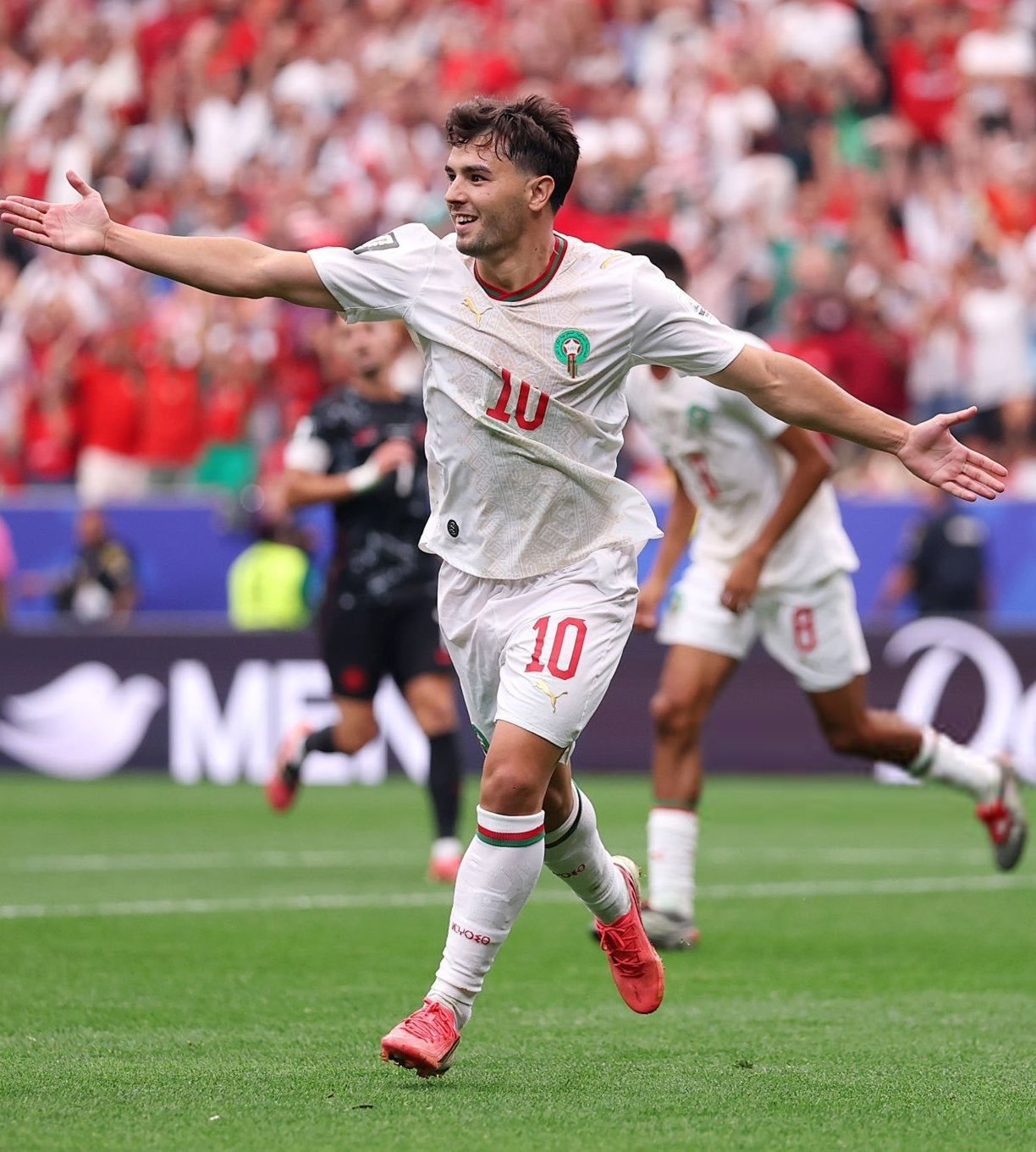 Soccer player in a white kit with number 10 celebrates a goal with arms outstretched on the field.