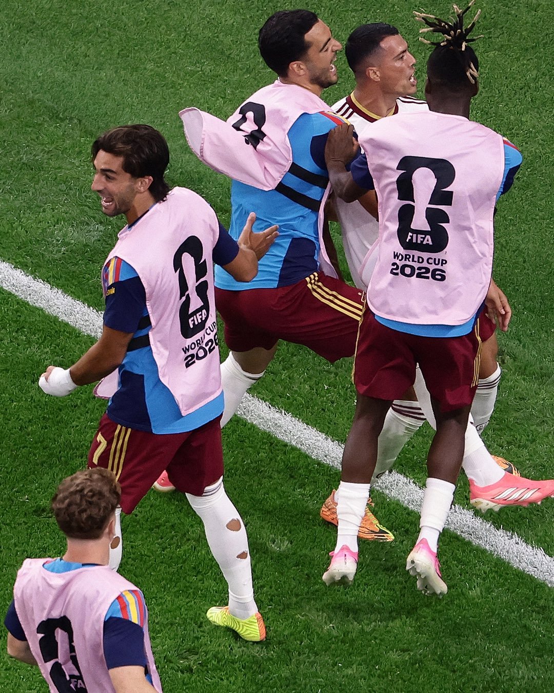 Soccer players in pink training bibs celebrate on a green pitch, with FIFA World Cup 2026 logos visible on their backs. Soccer players in pink training bibs celebrate on a green pitch, with FIFA World Cup 2026 logos visible on their backs.
