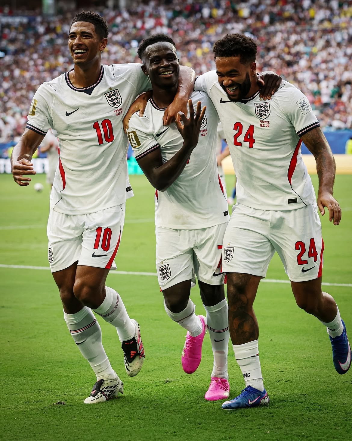 Three England footballers in white kit celebrate a goal with arms around each other on a green pitch, crowd in the background (numbers 10 and 24 visible). Three England footballers in white kit celebrate a goal with arms around each other on a green pitch, crowd in the background (numbers 10 and 24 visible).