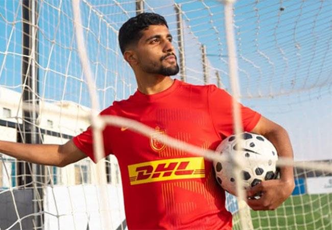 Male soccer player in a red DHL jersey leans on a goal net, holding a soccer ball under his arm on a sunny day. Male soccer player in a red DHL jersey leans on a goal net, holding a soccer ball under his arm on a sunny day.