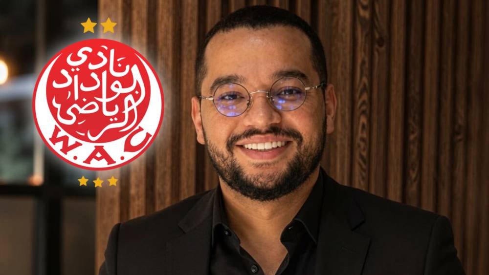 Smiling man in a black suit and glasses posed beside a red circular logo with Arabic script and gold stars on a wooden background (brand/award image).