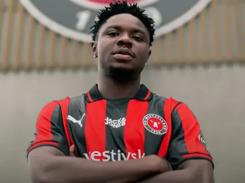Man wearing a red and black striped soccer jersey with arms crossed, standing in front of a stadium or sports arena background.