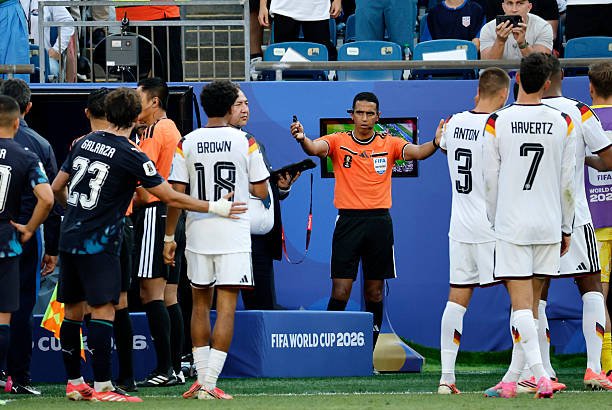 Soccer referee in an orange shirt raises his arms as players from two teams line up for kickoff before a FIFA World Cup 2026 match, with blue advertising boards and spectators in the stands behind them. Soccer referee in an orange shirt raises his arms as players from two teams line up for kickoff before a FIFA World Cup 2026 match, with blue advertising boards and spectators in the stands behind them.