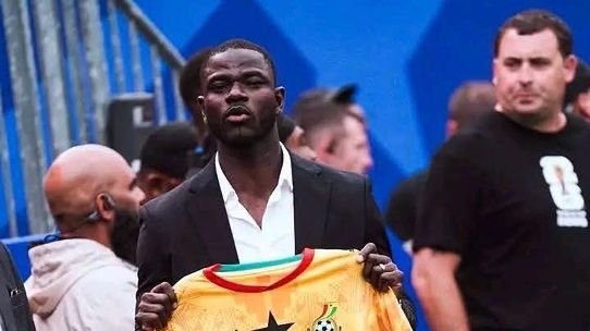 Man in a dark suit holding up a yellow jersey with a star, at an event with onlookers in the background.