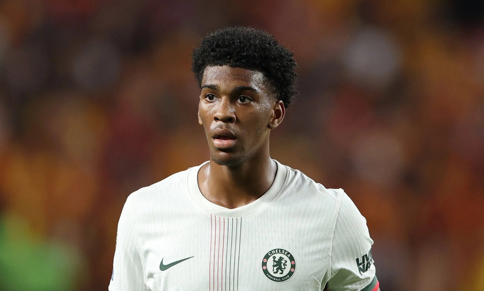 Chelsea footballer wearing a light gray jersey with a Chelsea crest, standing on a blurred stadium background, looking focused and sweaty from a match.