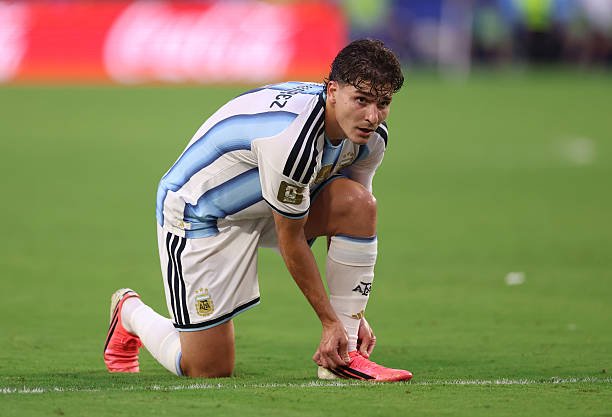Argentine soccer player in light-blue and white kit kneels on the grass, tying a bright pink cleat.
