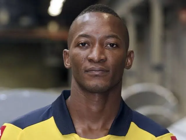 Portrait of a man with short hair wearing a yellow and navy polo, looking at the camera in a workshop setting