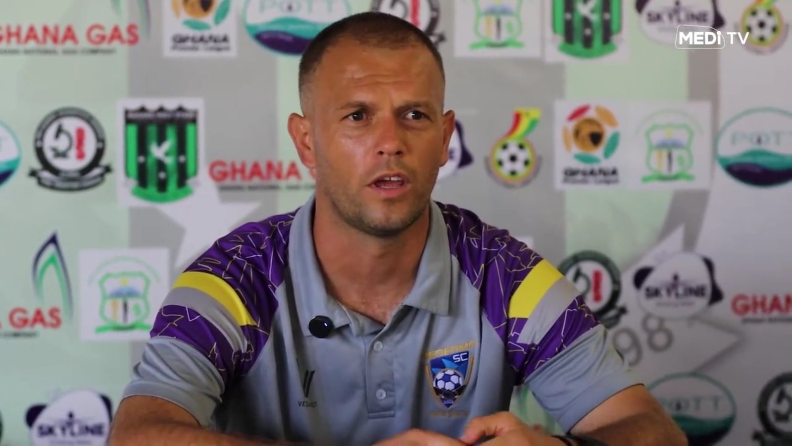 Man at a press conference wearing a gray polo with a crest, speaking into a mic with sponsor logos behind him. Man at a press conference wearing a gray polo with a crest, speaking into a mic with sponsor logos behind him.