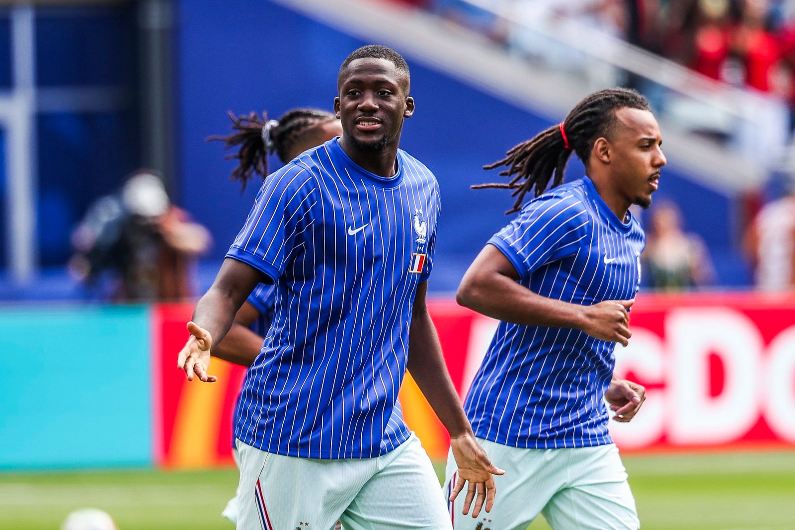 Two French national team players sprint on the soccer field in blue striped jerseys during a match, focused on the ball ahead of them.