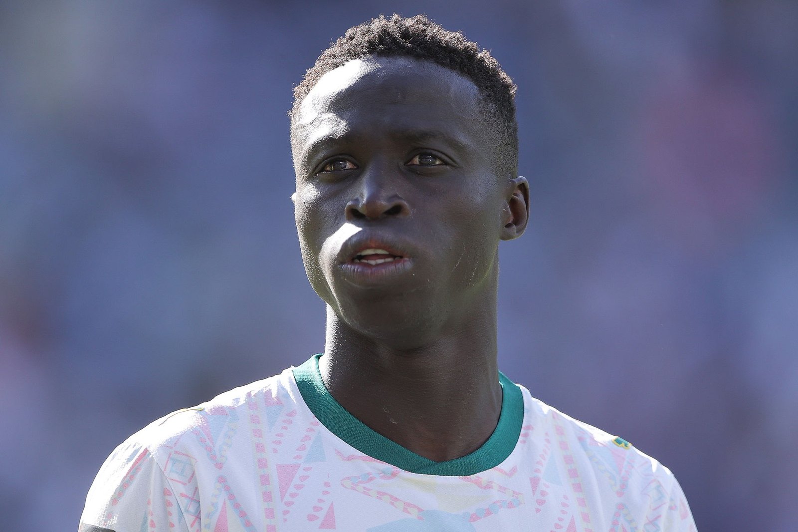 Close-up portrait of a Black male soccer player on the field, wearing a white jersey with teal trim, looking off-camera.