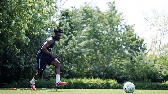 Athlete in a dark jersey runs toward a soccer ball on a grassy training field with trees in the background. Athlete in a dark jersey runs toward a soccer ball on a grassy training field with trees in the background.