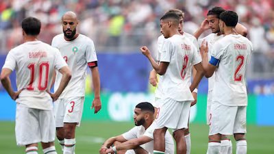 Group of soccer players in white uniforms with red numbers on a grass field, some standing and one kneeling, mid-game discussion.