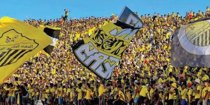 Large stadium crowd of yellow-clad fans waving flags and banners in support of their team.