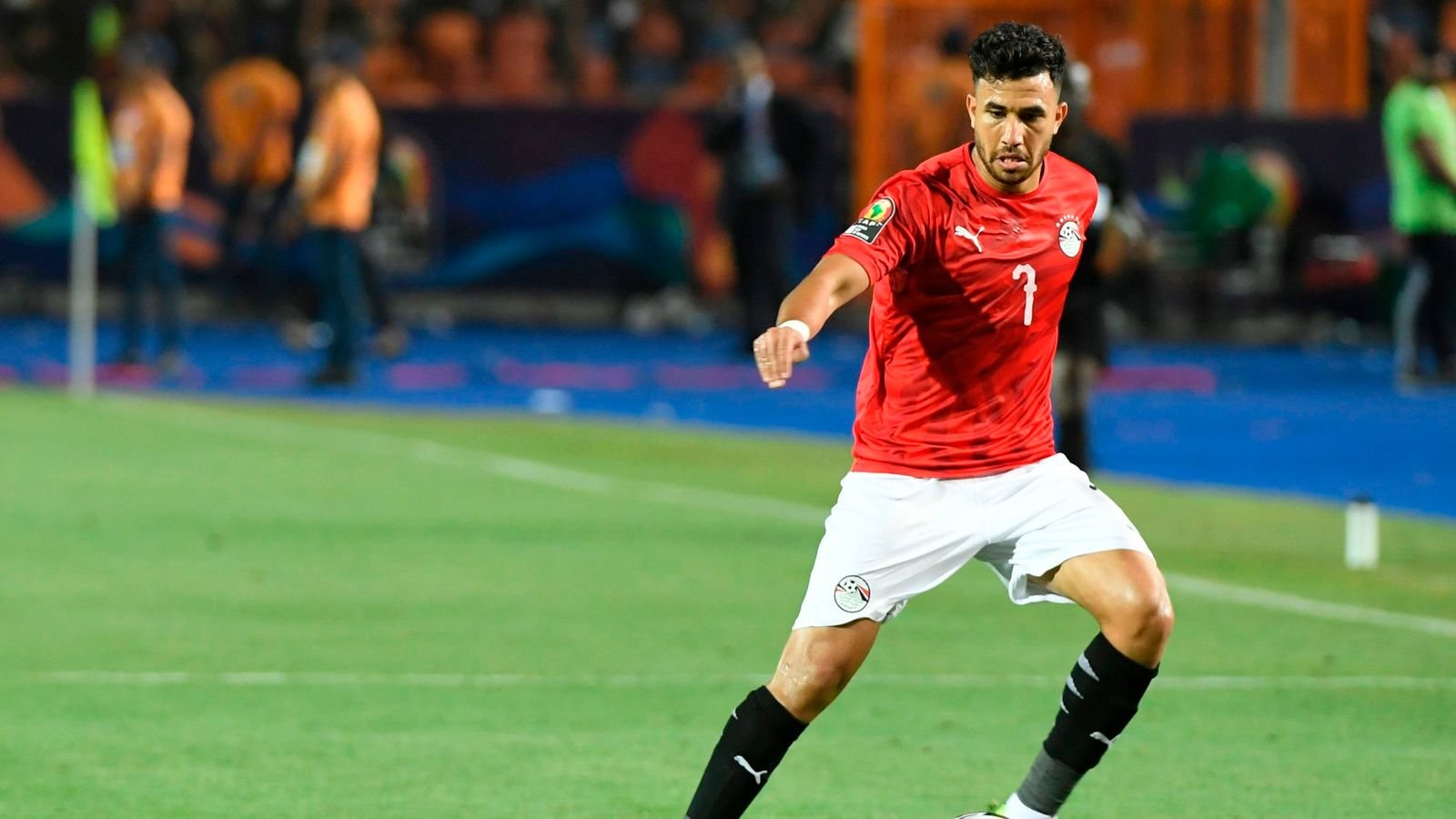 Soccer player in a red jersey (#7) dribbling on a green field during a match, with a stadium in the background. Soccer player in a red jersey (#7) dribbling on a green field during a match, with a stadium in the background.