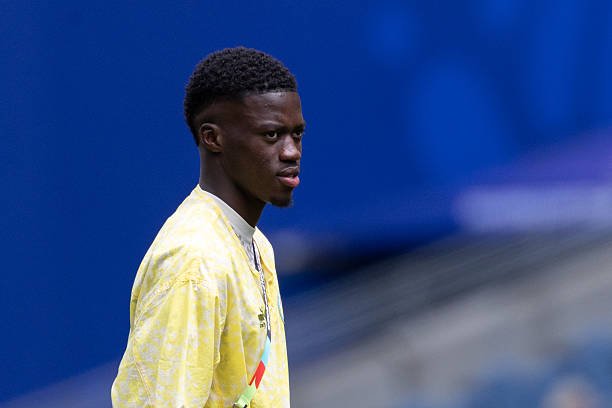 Young man in a pale yellow shirt standing outdoors against a blue background, looking to the left.