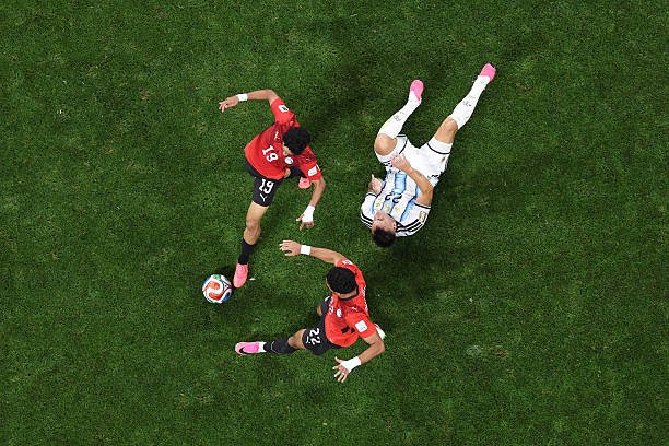 Aerial view of three soccer players contesting the ball on a green field: two in red jerseys and one in a white-and-blue striped kit, all mid-action. Aerial view of three soccer players contesting the ball on a green field: two in red jerseys and one in a white-and-blue striped kit, all mid-action.