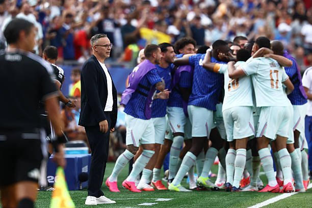 Soccer players in light-blue kit celebrate in a huddle on the field as a coach watches nearby. Soccer players in light-blue kit celebrate in a huddle on the field as a coach watches nearby.