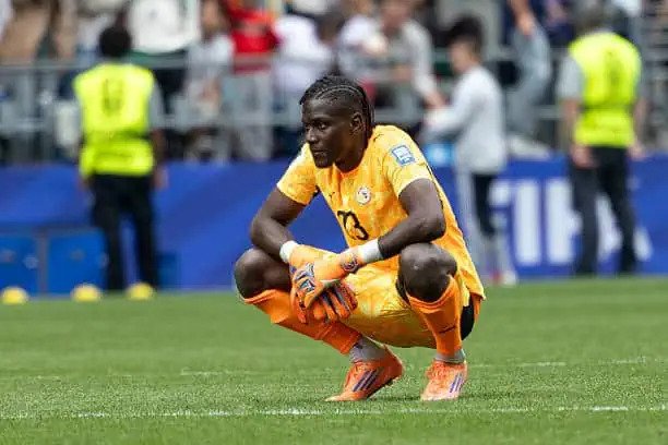 Soccer goalkeeper in an orange kit crouching on the pitch wearing gloves during a match.