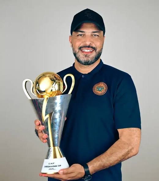 Smiling man in a dark polo and cap holds a tall silver trophy with a gold soccer ball on top.