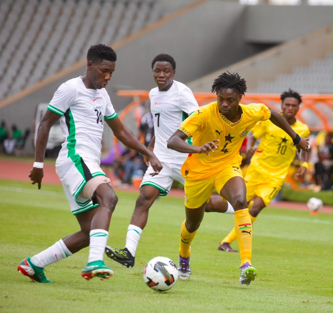 Soccer players in white and green and a yellow kit race for the ball on a grassy pitch during a match.
