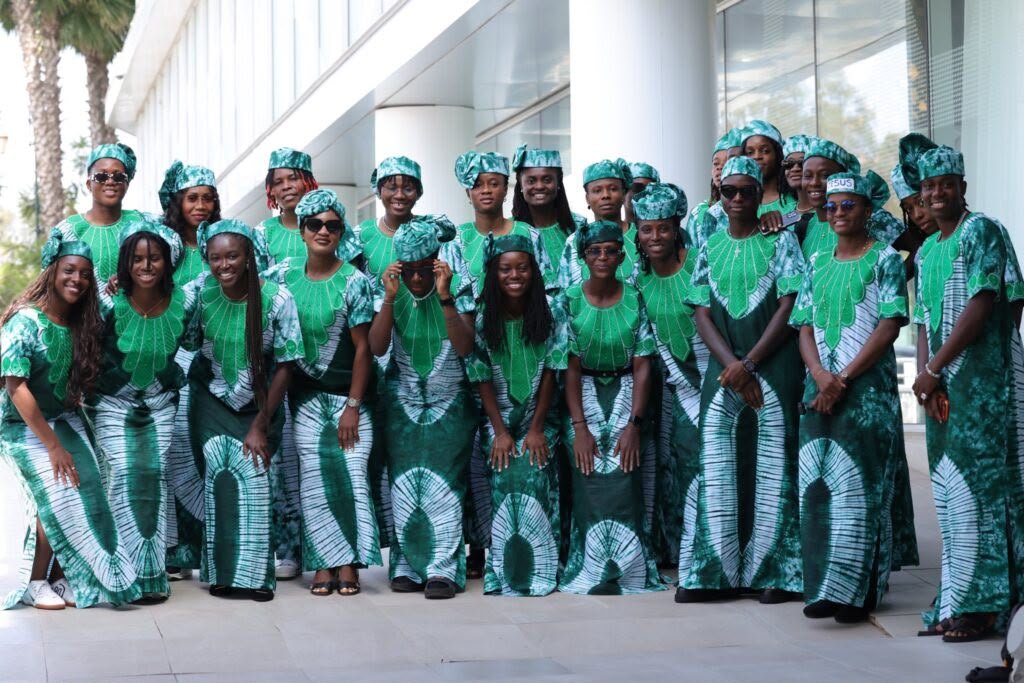 Group of people in matching green and white traditional outfits posing outside a glass-fronted building in daylight.