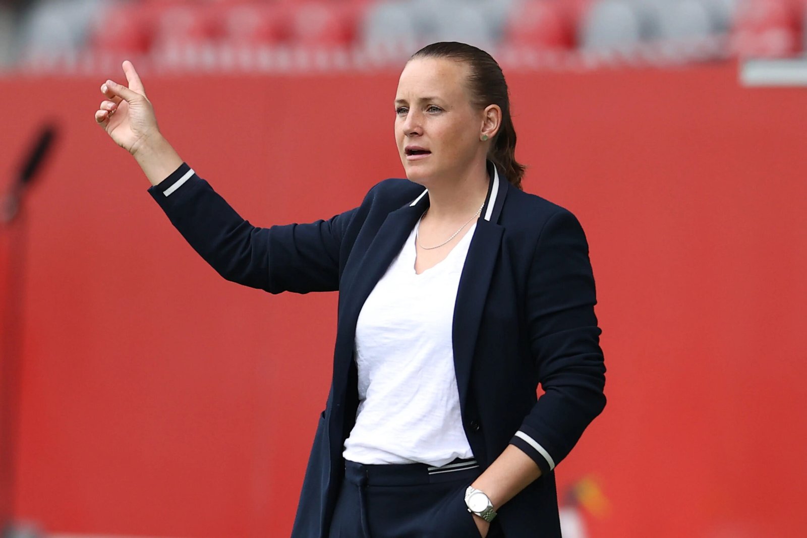 Woman in a navy blazer and white T‑shirt raising her right hand while speaking, in front of a red stadium backdrop