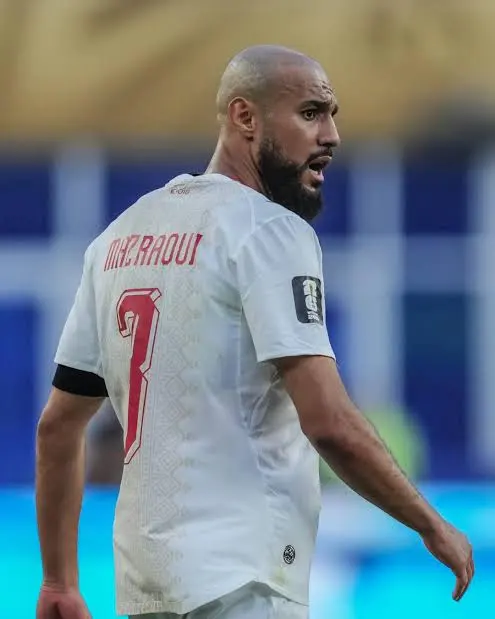 Soccer player in a white jersey with the number 3 and name on the back, looking to the right; wearing a black armband on his left arm on the field.