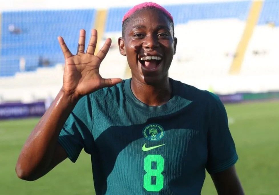 Soccer player with pink hair in a teal jersey (number 8) waving and smiling on a sunny field, stadium seating in the background.