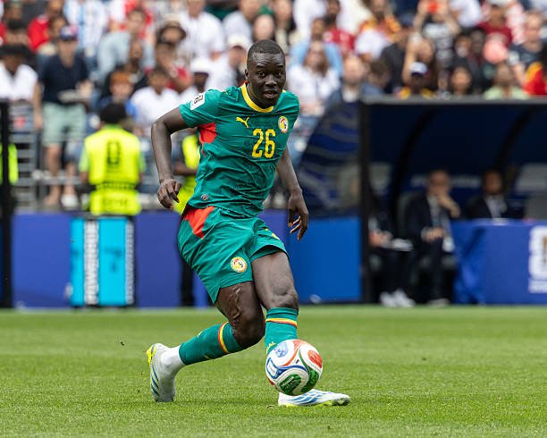 Soccer player in teal jersey number 26 dribbles a ball on a grass field during a match, with spectators in the stands behind him. Soccer player in teal jersey number 26 dribbles a ball on a grass field during a match, with spectators in the stands behind him.